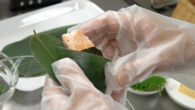 Maki places salt-grilled cherry salmon onto an onigiri, and secures it with nori. To finish, she wraps it in a bamboo leaf. Mr. Takayanagi and his wife help out, too.